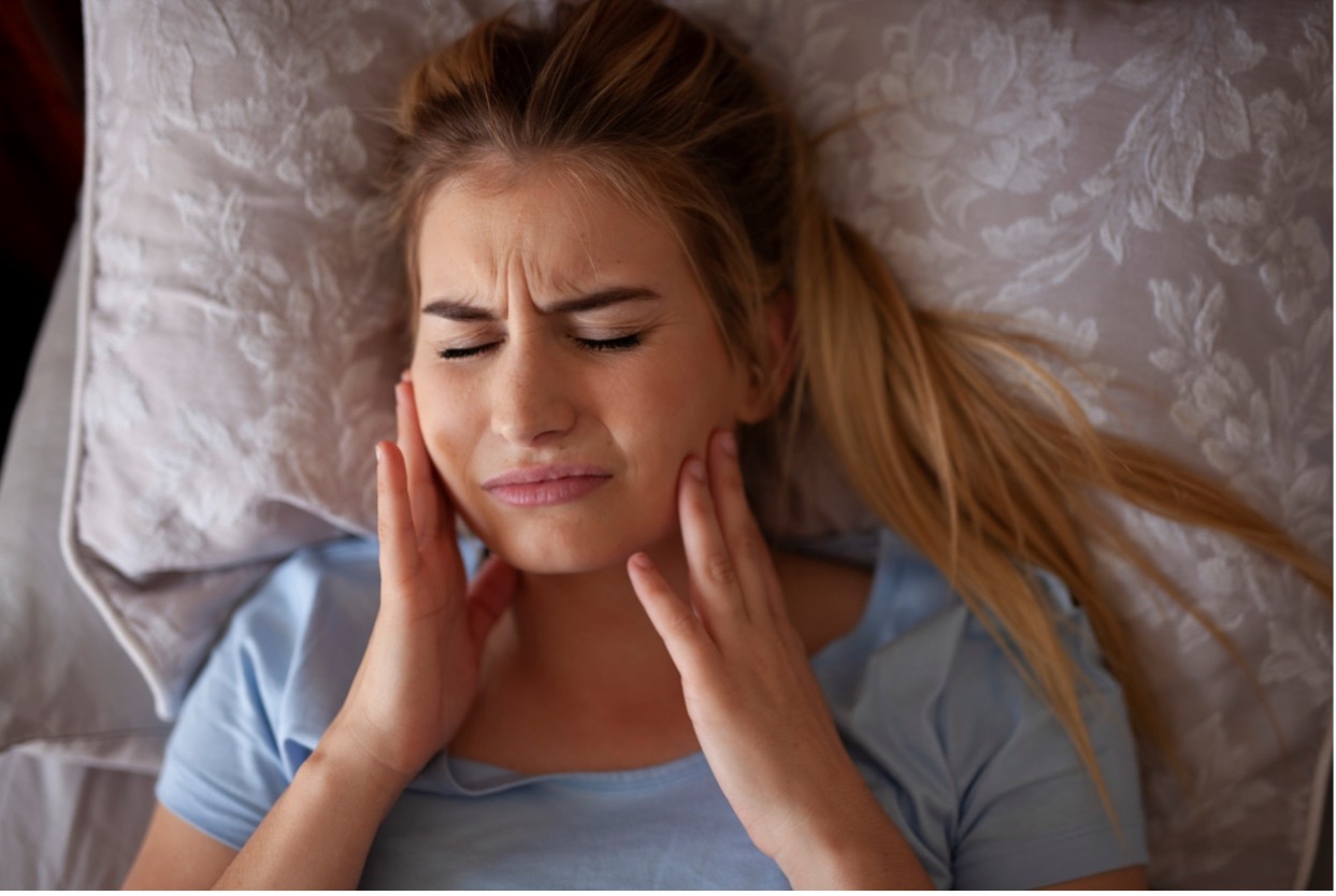 person lying on bed holding their jaw, TMJ