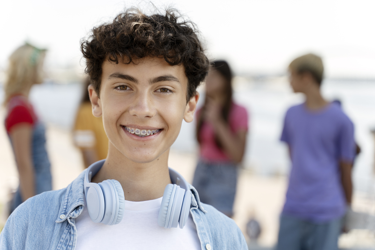 Portrait of smiling teenager with braces wearing headphones looking at camera standing on the street with friends on background. Summer concept, Getting Started with Braces or Clear Aligners: Your Summer Smile Transformation