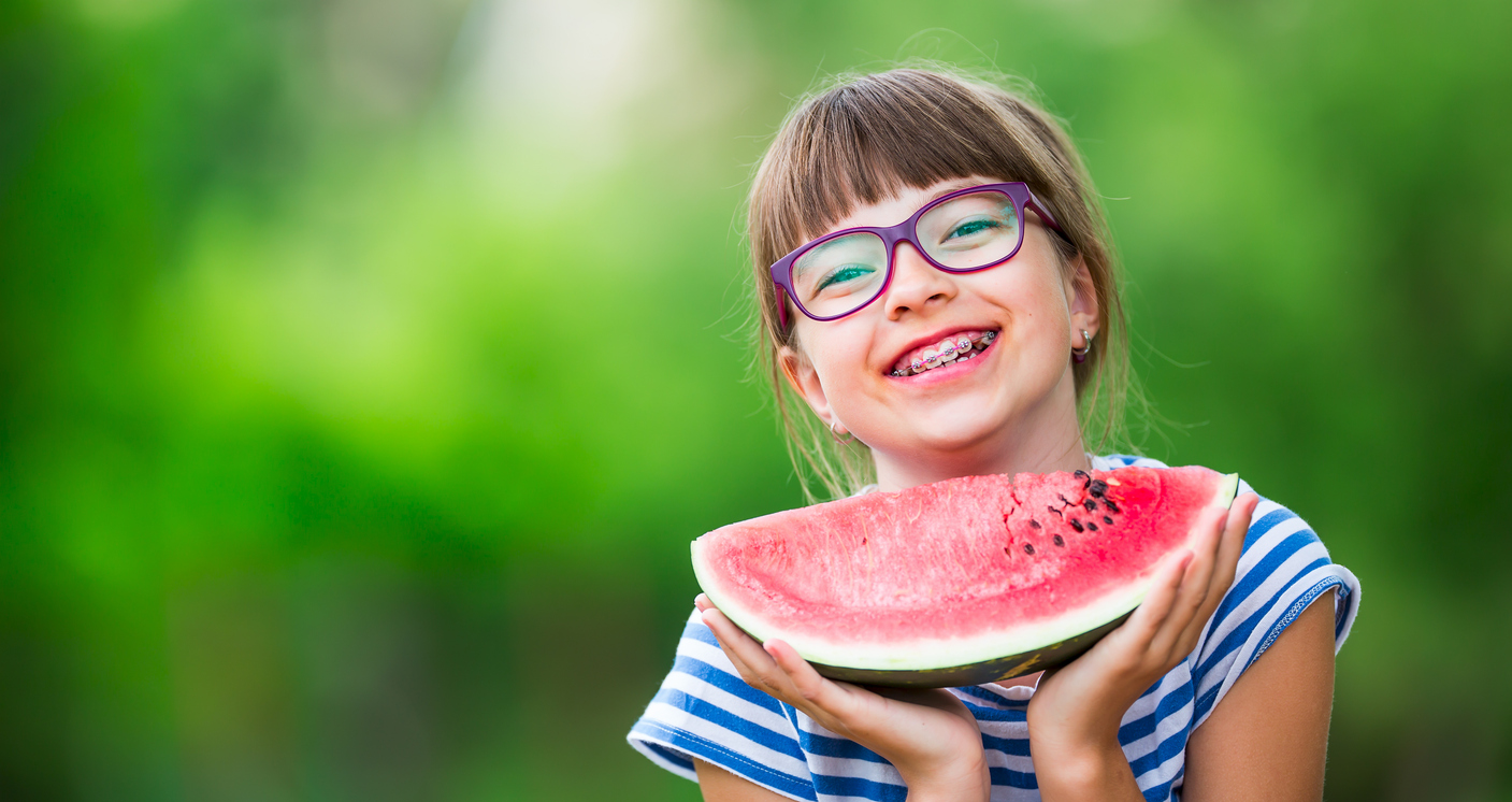 Child eating watermelon. Kids eat fruits in the garden. Pre teen girl in the garden holding a slice of water melon. happy girl kid eating watermelon. Girl kid with gasses and teeth braces.Child eating watermelon. Kids eat fruits in the garden. Pre teen girl in the garden holding a slice of water melon. happy girl kid eating watermelon. Girl kid with gasses and teeth braces.Summer Fun with Braces: Enjoying the Season with a Radiant Smile