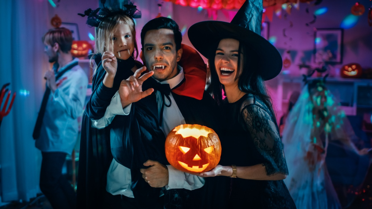 Family in Halloween costumes, including vampires and a witch, holding a carved pumpkin at a party.